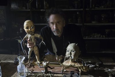 A man with messy hair looks forward behind a table covered in various handmade, eerie dolls and tools.
