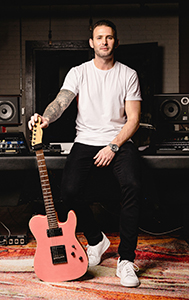 A man in a white t-shirt and black pants sits in a studio holding a pink electric guitar.
