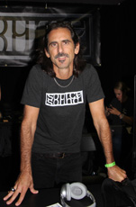 A man with long dark hair and a mustache wearing a black t-shirt with a white graphic stands behind a table with headphones.