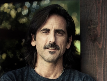 A man with dark, shoulder-length hair and a mustache looks toward the camera against a blurred background.