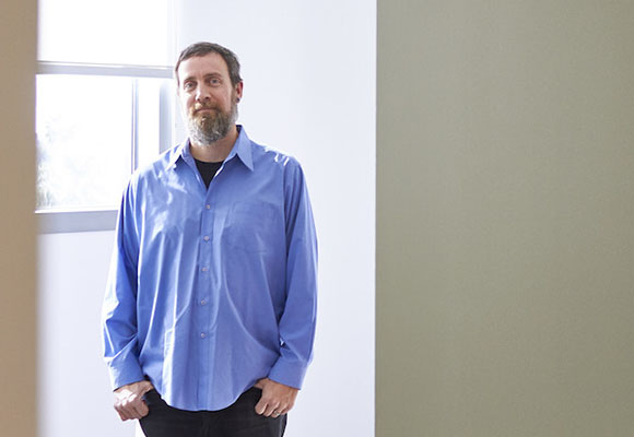 A bearded man wearing a blue button-down shirt stands in a doorway with his hands in his pockets.