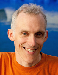 A man with short graying hair and a wide smile wearing an orange t-shirt against a blue background.