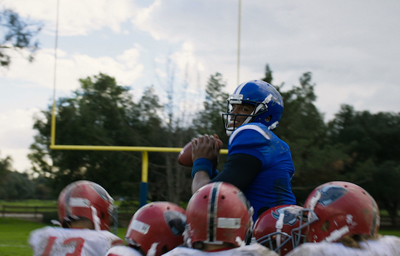 A football player in a blue jersey and helmet holds a football, surrounded by teammates in red helmets.