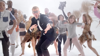 A man in a black tuxedo and glasses dances in the center of a group of people in diverse costumes.