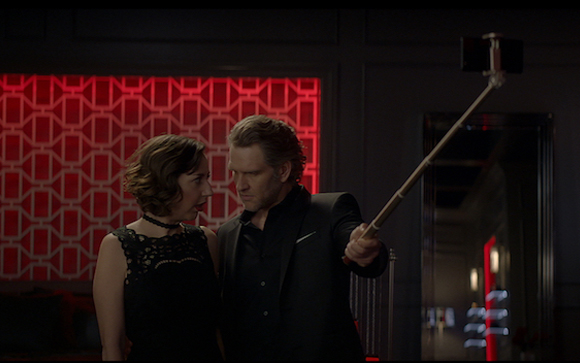 A man in a black suit and a woman in a black lace dress pose for a selfie using a selfie stick.