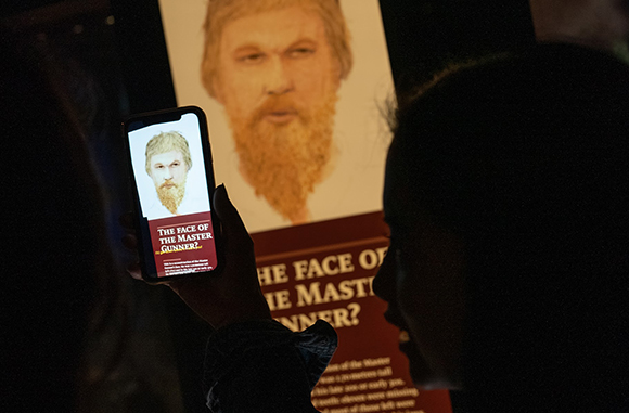 A person holds a smartphone showing a portrait of a bearded man, positioned in front of a large museum display.