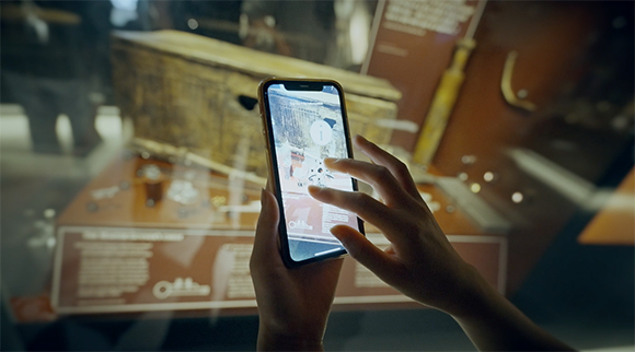 Hands holding a smartphone displaying an augmented reality view of a museum artifact behind glass.