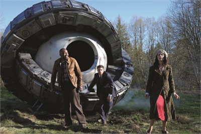 A man in a brown jacket, a man in a suit, and a woman in a long coat stand before a massive metal sphere.