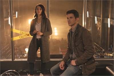 A woman in a long cardigan stands by a window while a man in a brown jacket sits in the foreground.