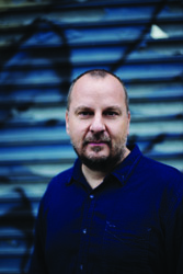 A man with short hair and a beard wearing a dark blue button-down shirt against a blue textured background.