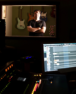 A man with crossed arms stands in a studio booth before several electric guitars.