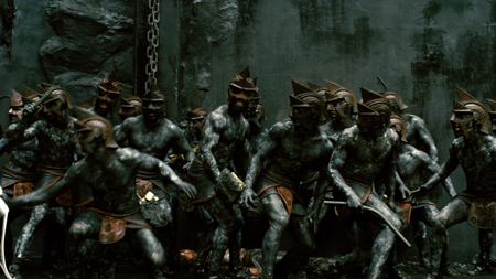 A group of muscular warriors in bronze helmets and loincloths standing in a dark, stone environment.