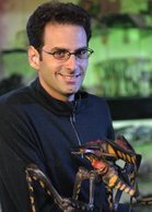 A man with glasses and dark hair smiles while holding a large, detailed insect sculpture.