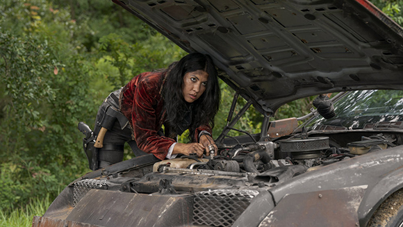 A woman in a red velvet jacket leans over an open car hood to inspect the engine.