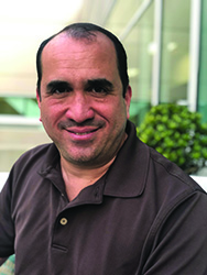 A man with dark hair and a brown polo shirt smiling at the camera.