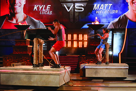 Two men pull on ropes attached to heavy pillars on separate raised platforms under studio lights.