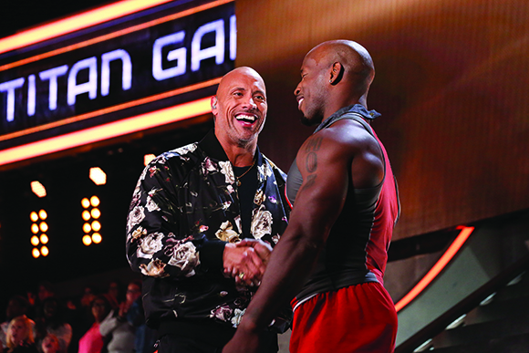Two muscular men smiling and shaking hands in front of a large glowing sign.