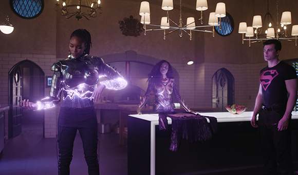 A woman in glowing purple armor stands in a kitchen while a man in a Superman shirt watches.
