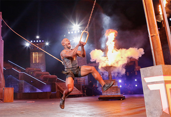 A muscular man swings on a trapeze during a nighttime stage performance with fire and smoke.