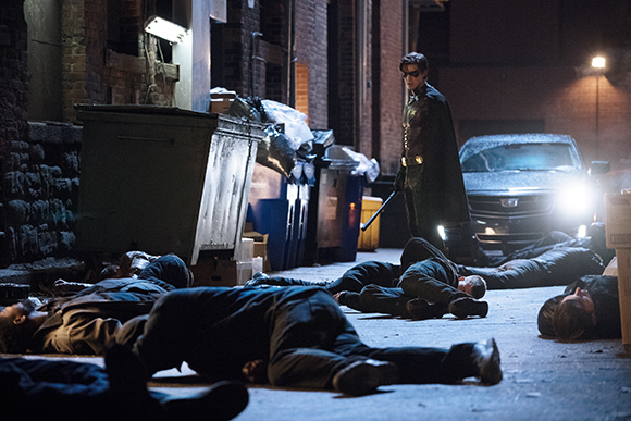 A person in a dark cape and mask stands in a dark alley next to several people lying on the ground.