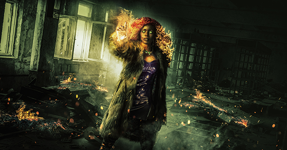A woman with glowing orange hair and a hand emitting fire stands amidst debris in a dark, ruined room.