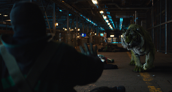 A large tiger walks toward a person wearing a dark hood in a dimly lit, industrial warehouse.