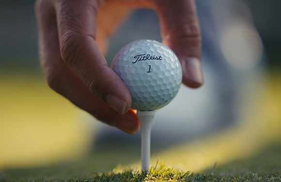 A hand places a white Titleist golf ball onto a white tee in the grass.