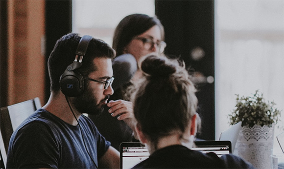 Three people sit at a desk with laptops and headphones in a bright office.