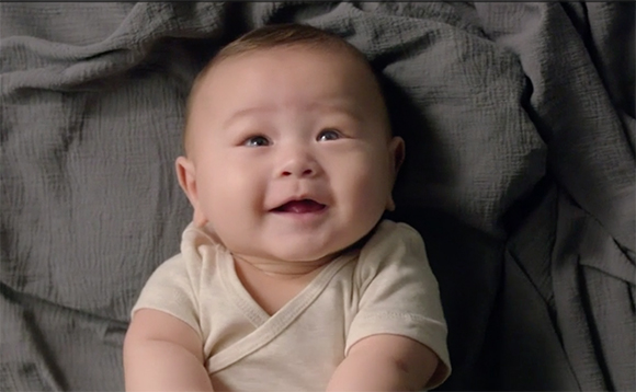 A smiling baby in a cream-colored shirt lying on a textured grey blanket.