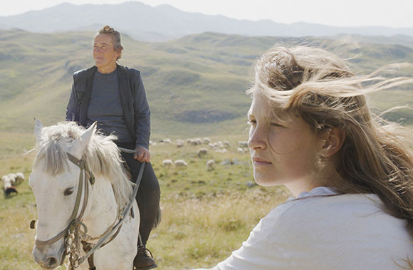 A woman rides a white horse in a grassy field while a young woman looks off-camera in the foreground.