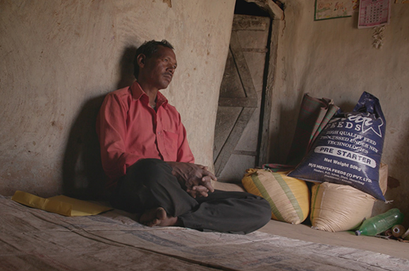 A man in a red shirt sits cross-legged on a floor next to large sacks of animal feed.