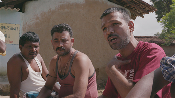 Three men sit together outdoors near a tan building under bright sunlight.