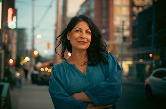 A woman with dark hair and a blue blouse smiles with her arms crossed on a city street at sunset.
