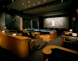 A darkened professional media screening room with tiered seating, control consoles, and a large projection screen.