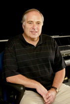 A man with short hair wearing a black striped polo shirt and khaki pants sits in a chair.