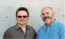 Two men, one wearing sunglasses and a dark shirt, the other with a beard and a blue shirt, standing together.