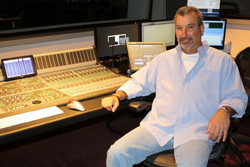 A man in a light blue shirt sits at a professional audio mixing desk surrounded by computer monitors.