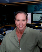 A man in a green collared shirt smiling in a studio with computer monitors in the background.