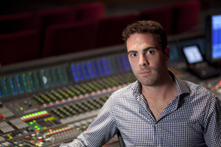 A man in a checkered shirt sits behind a large professional audio mixing console with many sliders and lights.