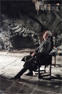 A man in dark clothing sits in a wooden chair before a large dragon relief carved into a stone wall.