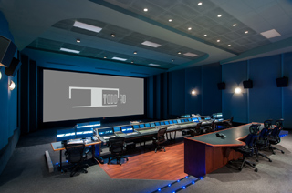 A dark media control room with a large projection screen and long rows of workstations.
