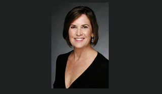 A smiling woman with short brown hair wearing a black V-neck top against a gray background.