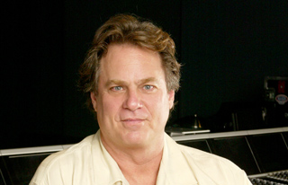 A man with wavy brown hair and blue eyes wearing a cream-colored button-down shirt.