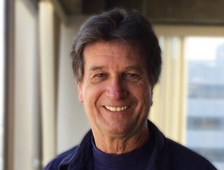 A middle-aged man with dark hair smiling at the camera against a blurred window background.