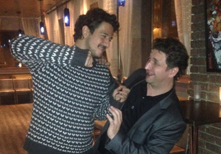 Two men are playfully sparring indoors. One man in a patterned sweater grabs the other man's neck while the second man holds