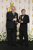 Two men in black tuxedos smiling and holding gold Oscar awards in front of a white backdrop.