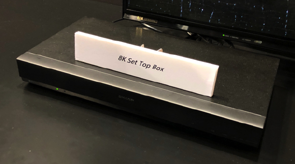 A black Sharp 8K Set Top Box with a white sign that reads "8K Set Top Box" is on a dark surface, with a television screen vis