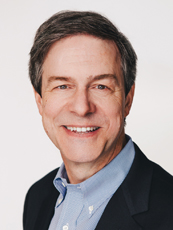A smiling middle-aged man with short brown hair wearing a light blue shirt and dark blazer.