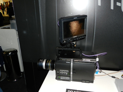 Professional video camera with lens and attached monitor, set on a white table.