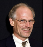 A man with short blond hair, glasses, and a mustache smiles. He is wearing a suit jacket over a collared shirt and tie.